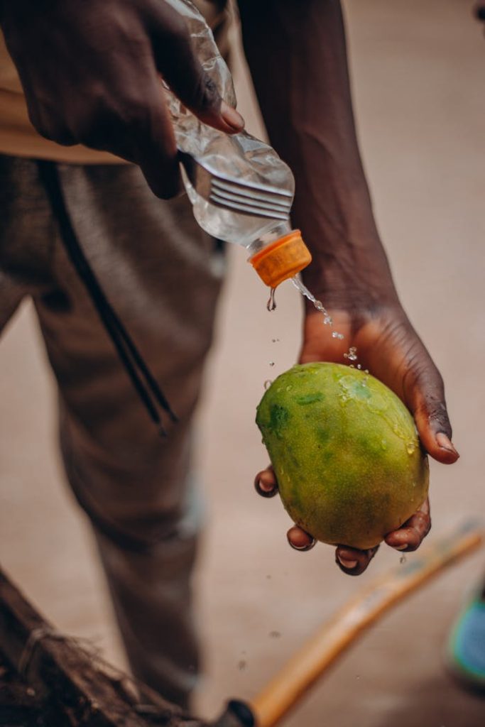 A person washing a ripe mango with bottled water outdoors, focusing on cleanliness and freshness.